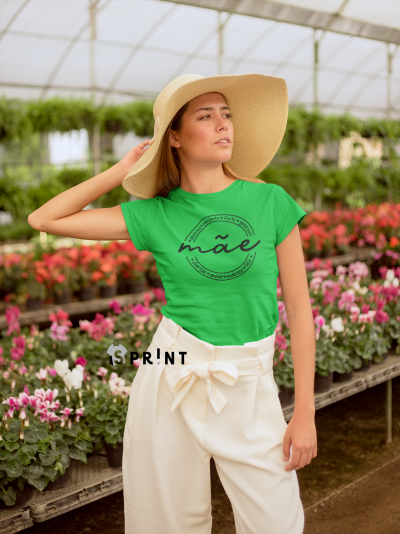 Mulher com t-shirt verde estampada, calças brancas largas e chapéu de palha, em estufa com flores