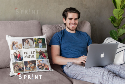 Almofada personalizada com fotos e texto FELIZ NATAL num sofá junto a homem com portátil.