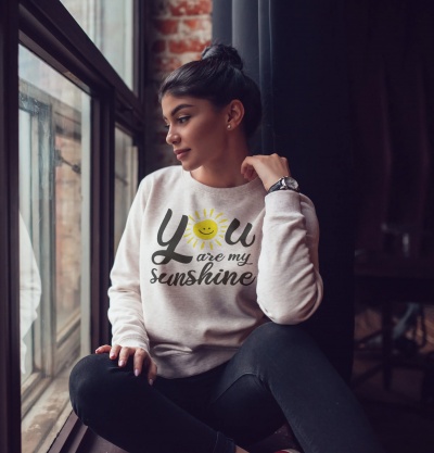 Mulher sentada junto a janela usando sweatshirt branca com estampa colorida de sol e texto