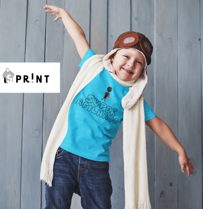 Menino com camisola azul 'SUPER AFILHINHO', calças de ganga, cachecol bege e chapéu de aviador castanho, fundo madeira cinzenta.