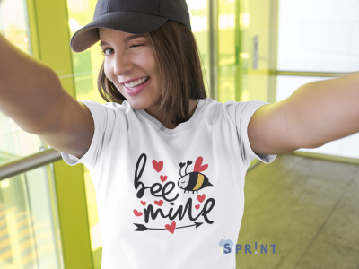 T-shirt branca com estampado 'bee mine' e abelha, usada por mulher com chapéu preto, em ambiente interior amarelo