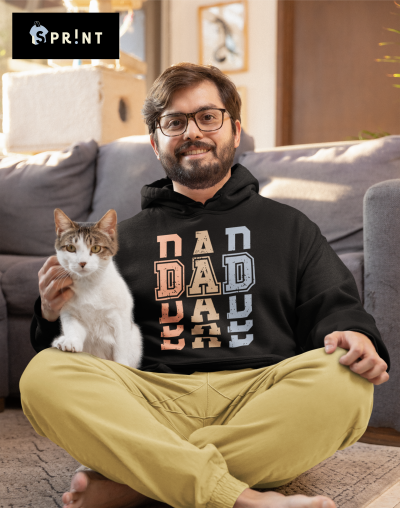 Homem sentado no chão com hoodie preto com letras 'DAD' coloridas e gato no colo
