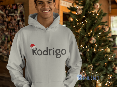 Homem a sorrir a usar camisola branca com capuz com nome 'Rodrigo' e gorro de Natal, árvore de Natal decorada atrás