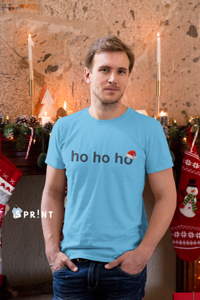 Homem com t-shirt azul clara com texto 'ho ho ho' e chapéu de Natal, ambiente natalício com lareira e decoração