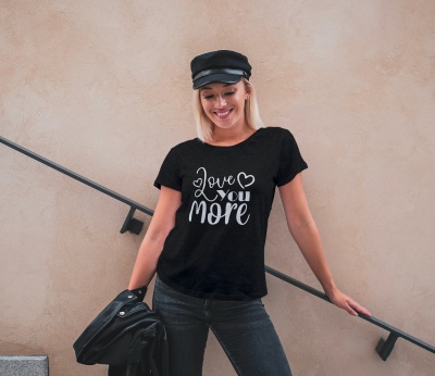 Mulher com t-shirt preta com texto branco 'love you more' e calças negras junto a parede bege