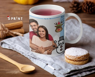 Caneca branca com fotografia de casal e desenhos natalícios, sobre pano cinza, com colher de madeira e doce ao lado.