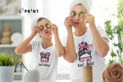 Mãe e filha vestindo t-shirts brancas com estampas semelhantes e texto 'tal mãe' e 'tal filha' em ambiente doméstico de banheiro.