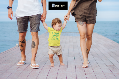 Criança com t-shirt amarela e calções bege segurando as mãos de dois adultos num passadiço junto ao mar