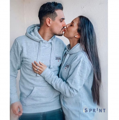 Casal usando hoodies cinzentos claros com inscrição Since 2015 e pequeno pássaro vermelho numa parede branca