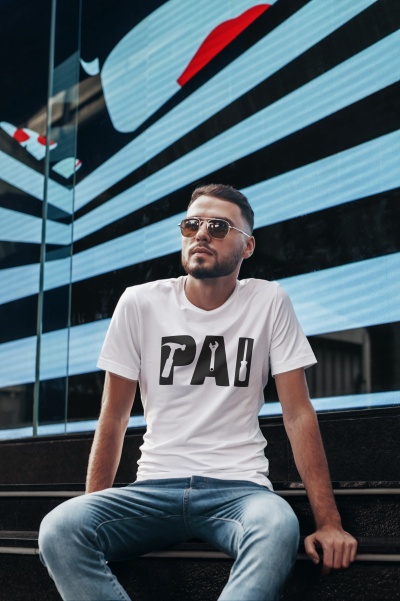 Homem com camisola branca 'PAI', jeans azuis e óculos de sol sentado