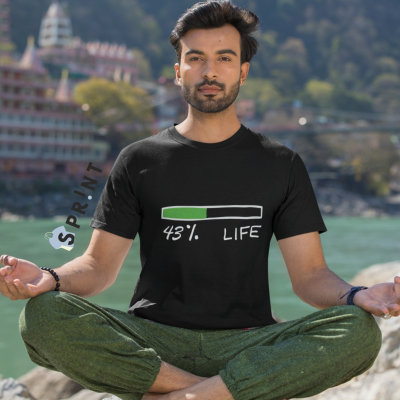 Homem a usar camisola preta com texto e barra de progresso, sentado junto a rio, com vegetação e edifícios ao fundo.