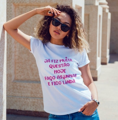 Mulher com t-shirt branca com texto em rosa e roxo, óculos escuros e cabelo encaracolado, em ambiente exterior com colunas de pedra.