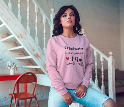 Mulher com sweatshirt rosa claro escrita Madrinha é segunda Mãe num ambiente interior com escada branca.