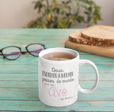 Caneca branca com texto em português sobre uma mesa de madeira verde clara, ao lado de óculos e pão.