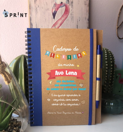 Caderno de receitas com capa decorada, elástico e caneta azul, sobre mesa com plantas e quadro flamingo ao fundo.