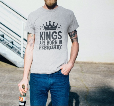 Homem com t-shirt cinza claro com estampa preta 'KINGS ARE BORN IN FEBRUARY' e calças jeans azuis, segurando garrafa ao ar livre