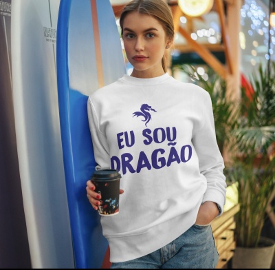 Mulher com sweatshirt branca estampada 'EU SOU DRAGÃO' segurando copo de café em ambiente com plantas
