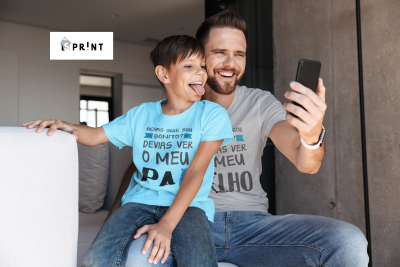 Homem e criança sentados num sofá cinza, usando t-shirts com texto em português, e o homem a tirar uma selfie com telemóvel