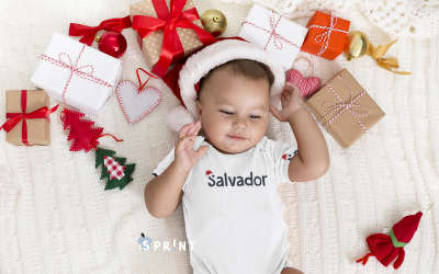 Bebé com body branco escrito Salvador e chapéu de Pai Natal rodeado de presentes e decorações natalinas