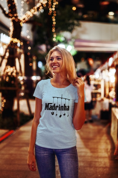 Mulher a sorrir usando t-shirt branca com texto 'Madrinha' num ambiente noturno iluminado