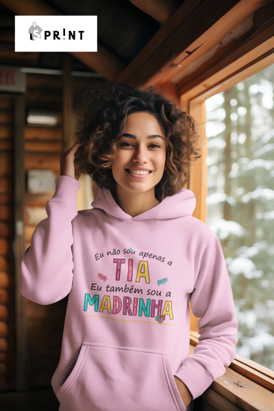 Mulher sorridente a usar suéter com capuz rosa claro com texto colorido, num ambiente de madeira junto a uma janela com neve lá fora