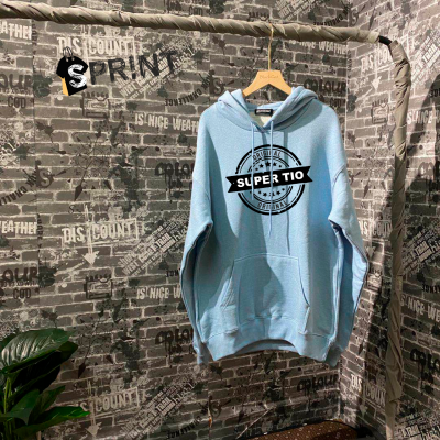 Sweatshirt azul clara com estampa SUPER TIO pendurada em cabide de madeira num cenário com parede cinza decorada com texto.