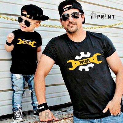 Pai e filho com t-shirts pretas estampadas com chave inglesa amarela e texto, usando bonés e óculos de sol, em frente a uma parede metálica.