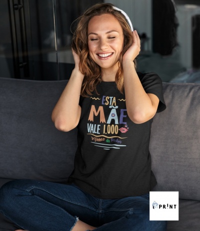 Mulher sorridente com camisola preta 'ESTA MÃE VALE 1.000 beijinhos do Pedro', sentada num sofá cinza escuro, calças de ganga e auscultadores brancos.