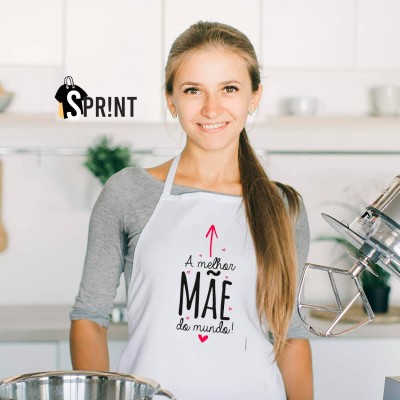 Mulher com avental branco com texto 'A melhor MÃE do mundo!' numa cozinha moderna, com misturador de massa e utensílios ao fundo.