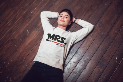 Mulher deitada em chão de madeira vestindo sweat cinzento com texto MRS. EST. 2020 e calças pretas