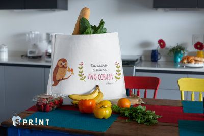 Sacola branca com estampa de coruja e texto sentado em mesa com frutas e vegetais