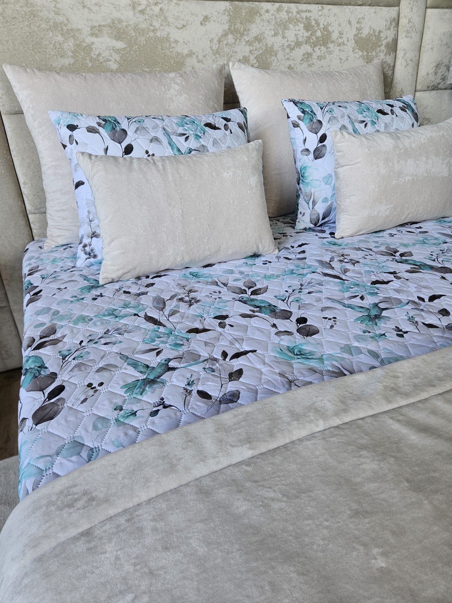 Cama com cabeceira estofada bege, colcha matelassê branca com flores azuis e almofadas bege e estampadas