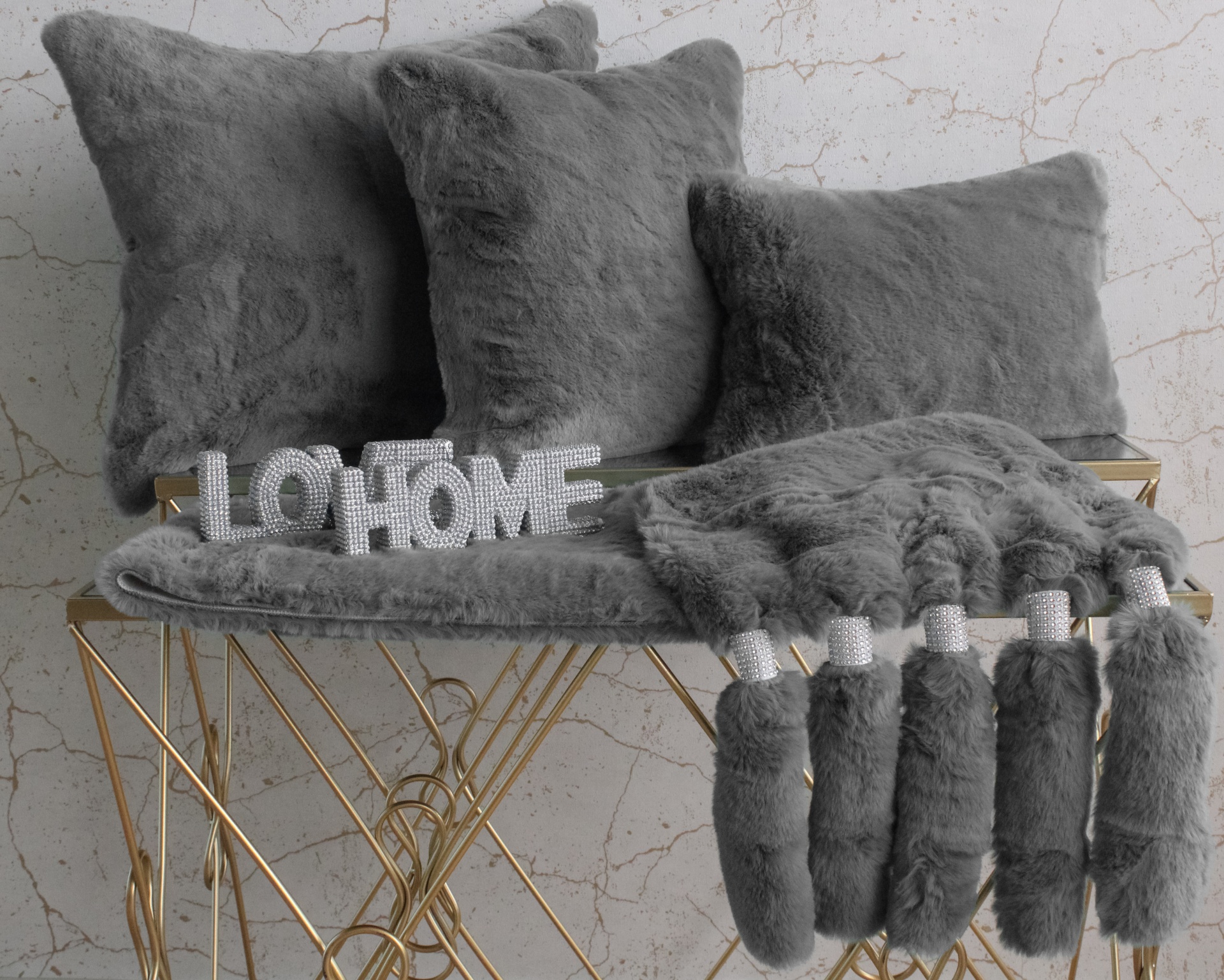 Almofadas e cobertor cinzentos de pelo sobre banco dourado com palavra decorativa LO HOME