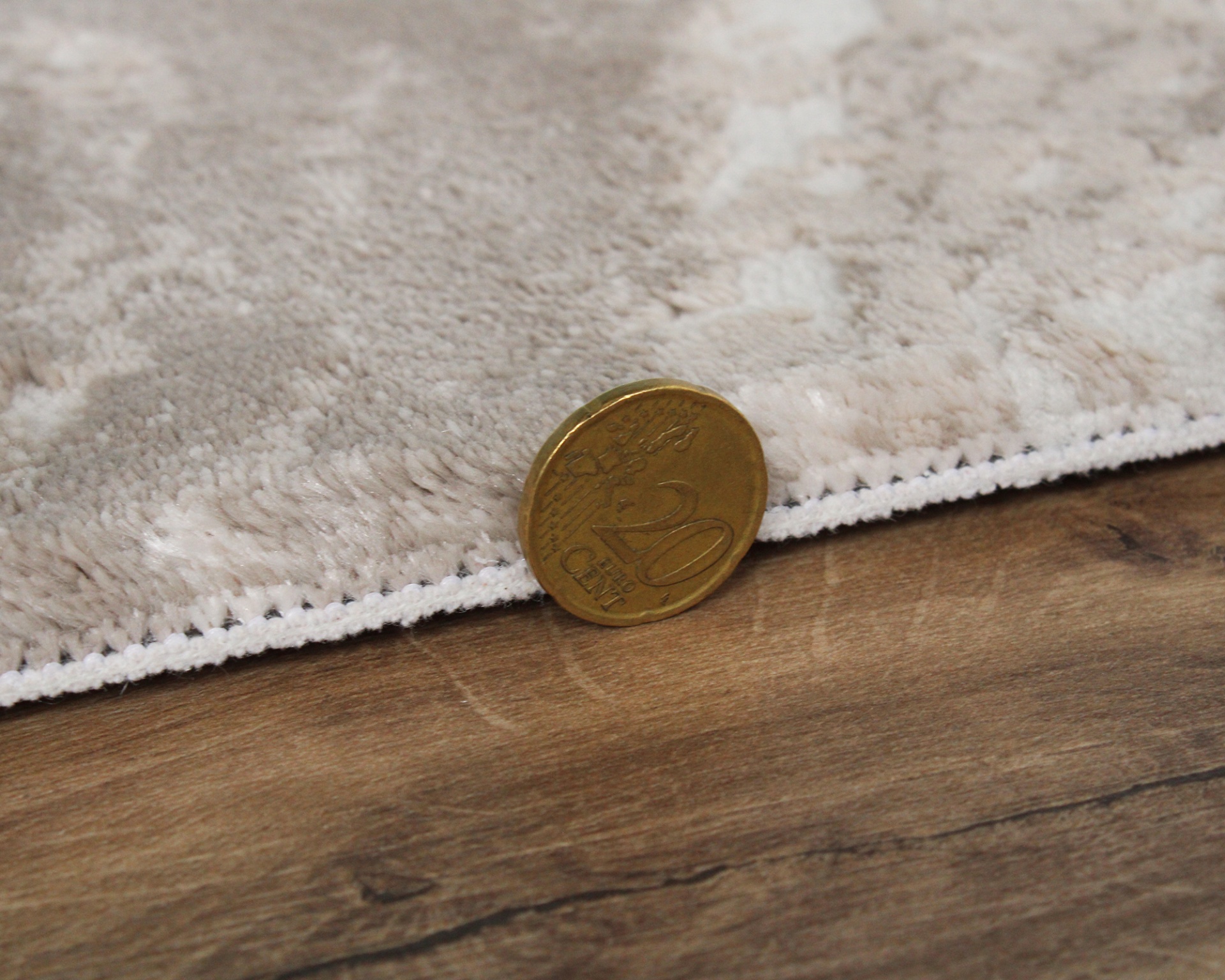 Moeda de 20 cêntimos junto a tapete bege com padrão floral em relevo sobre chão de madeira.