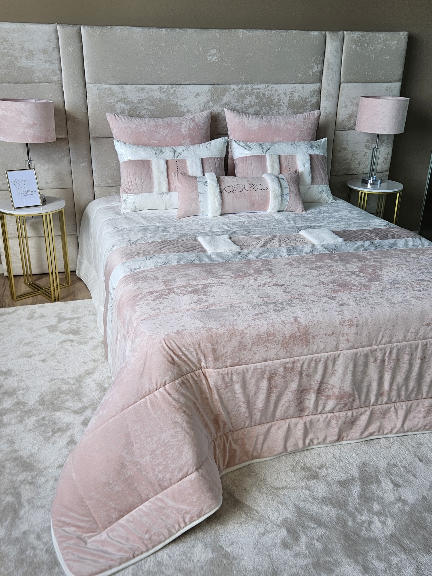 Edredão em Veludo Ref: Nepal Rosa Cama com roupa de cama rosa e bege aveludada e almofadas decorativas com padrão em retângulos, cabeceira estofada bege, mesas laterais douradas com tampo branco e candeeiros rosa.