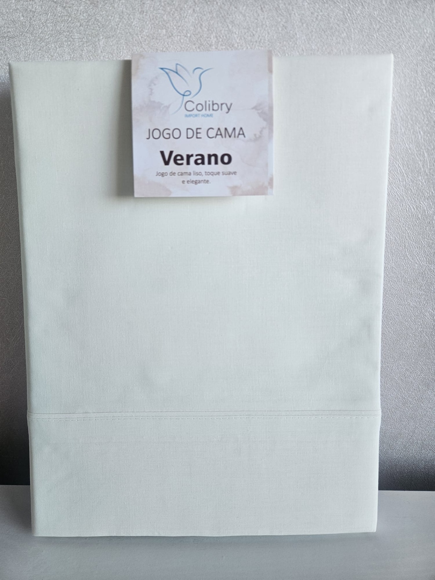 Jogo de cama branco com etiqueta Colibry Verano contra parede cinza