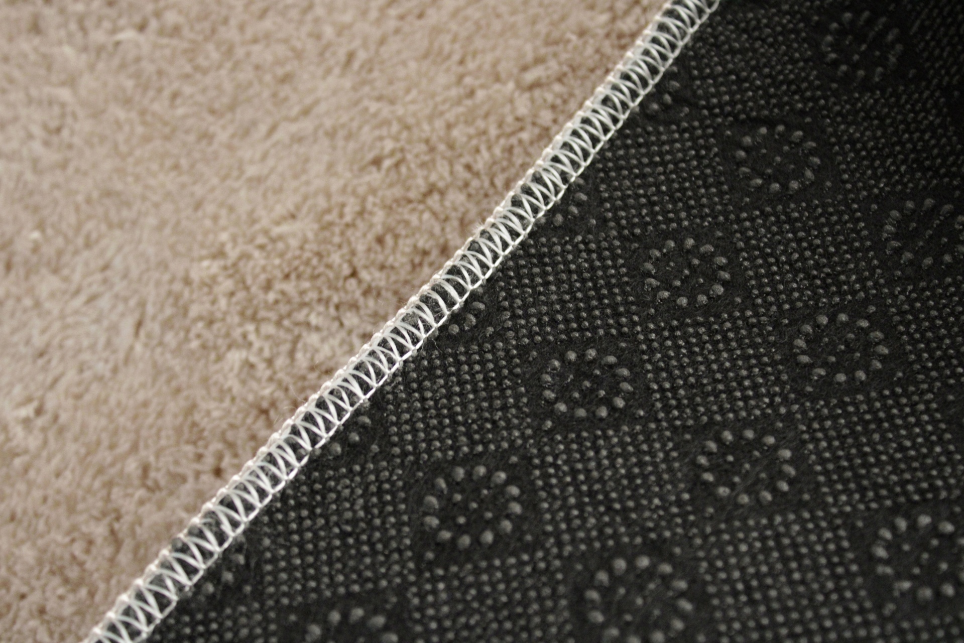 Detalhe da base antiderrapante de um tapete preto com padrões e costura branca ao lado de carpete bege