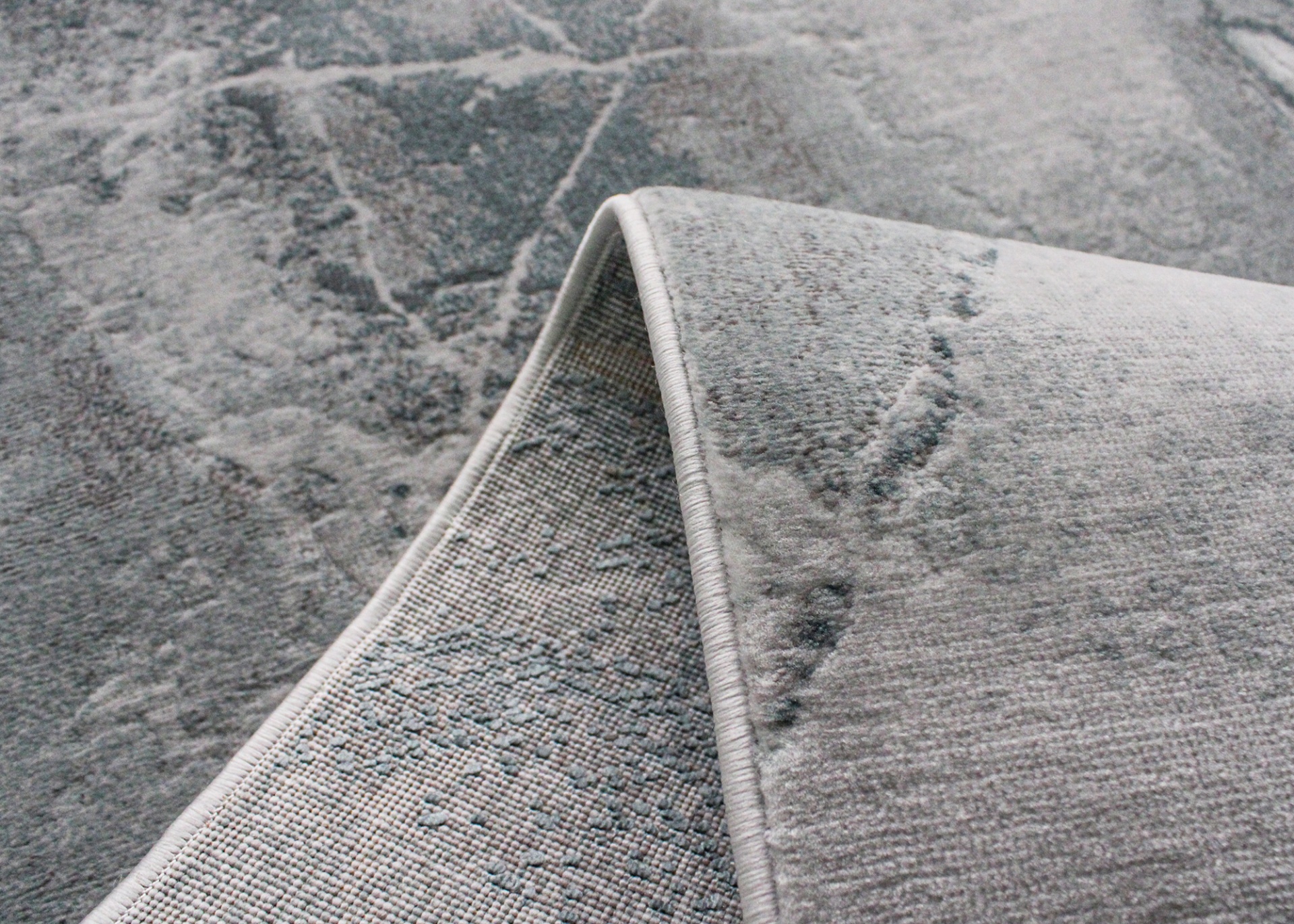 Tapete cinzento têxtil com padrão de fissuras e textura, dobrado para mostrar o verso.