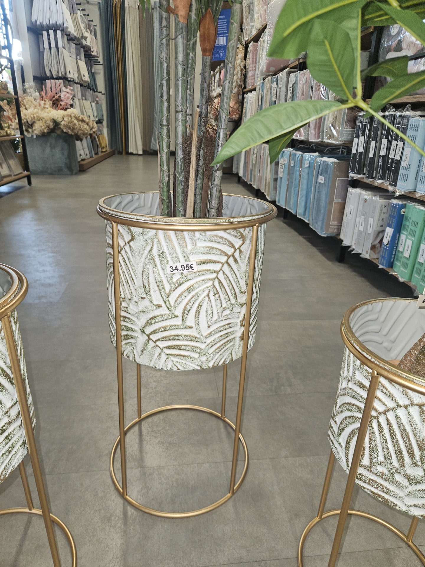 Vaso decorativo metálico dourado com padrão de folhas e preço 34.95€. Loja de decoração ao fundo.