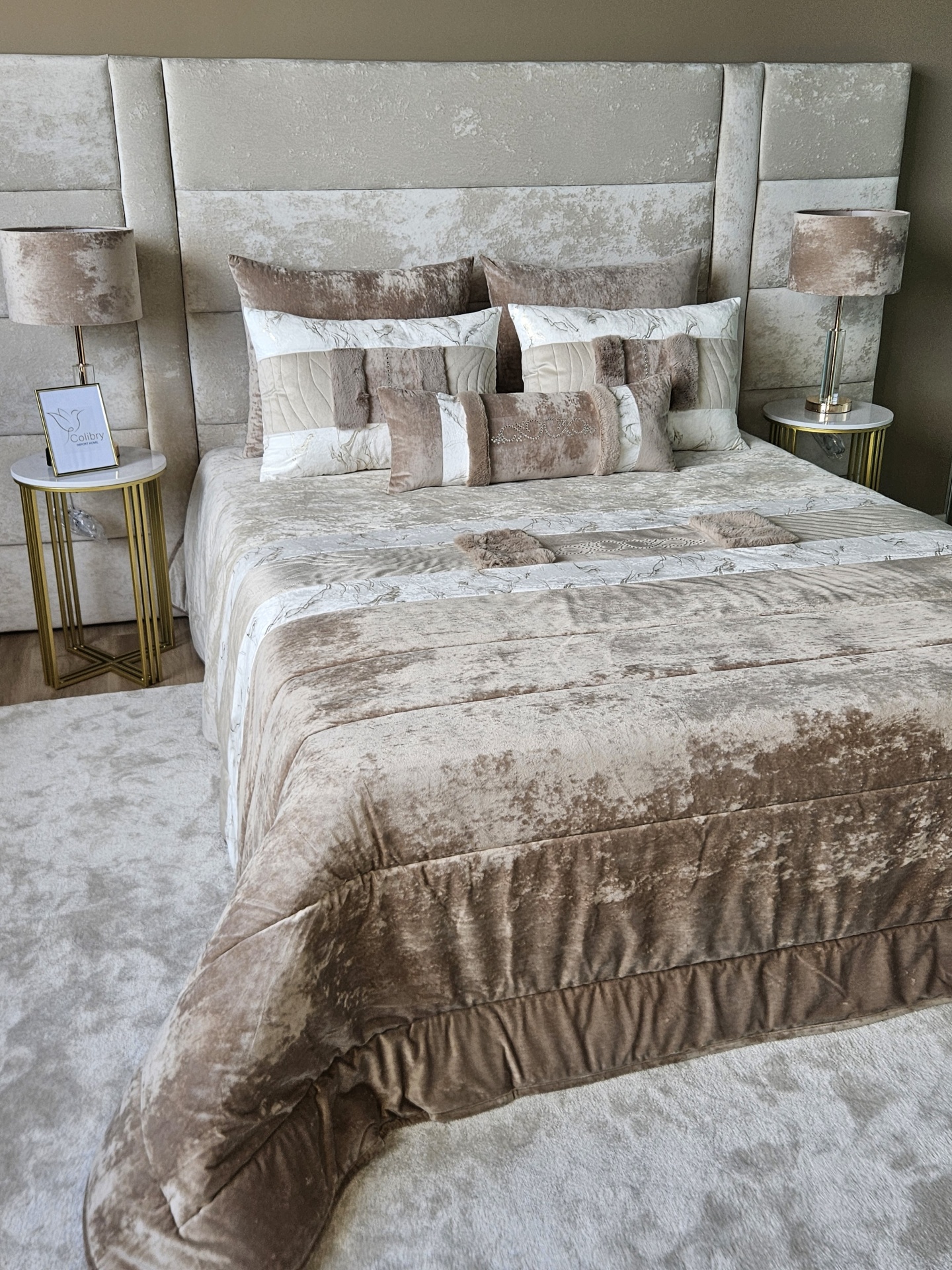 Edredão em Veludo Ref: Nepal Bege Cama estofada em veludo com roupa e almofadas em tons bege e creme, mesas de cabeceira douradas com candeeiros.
