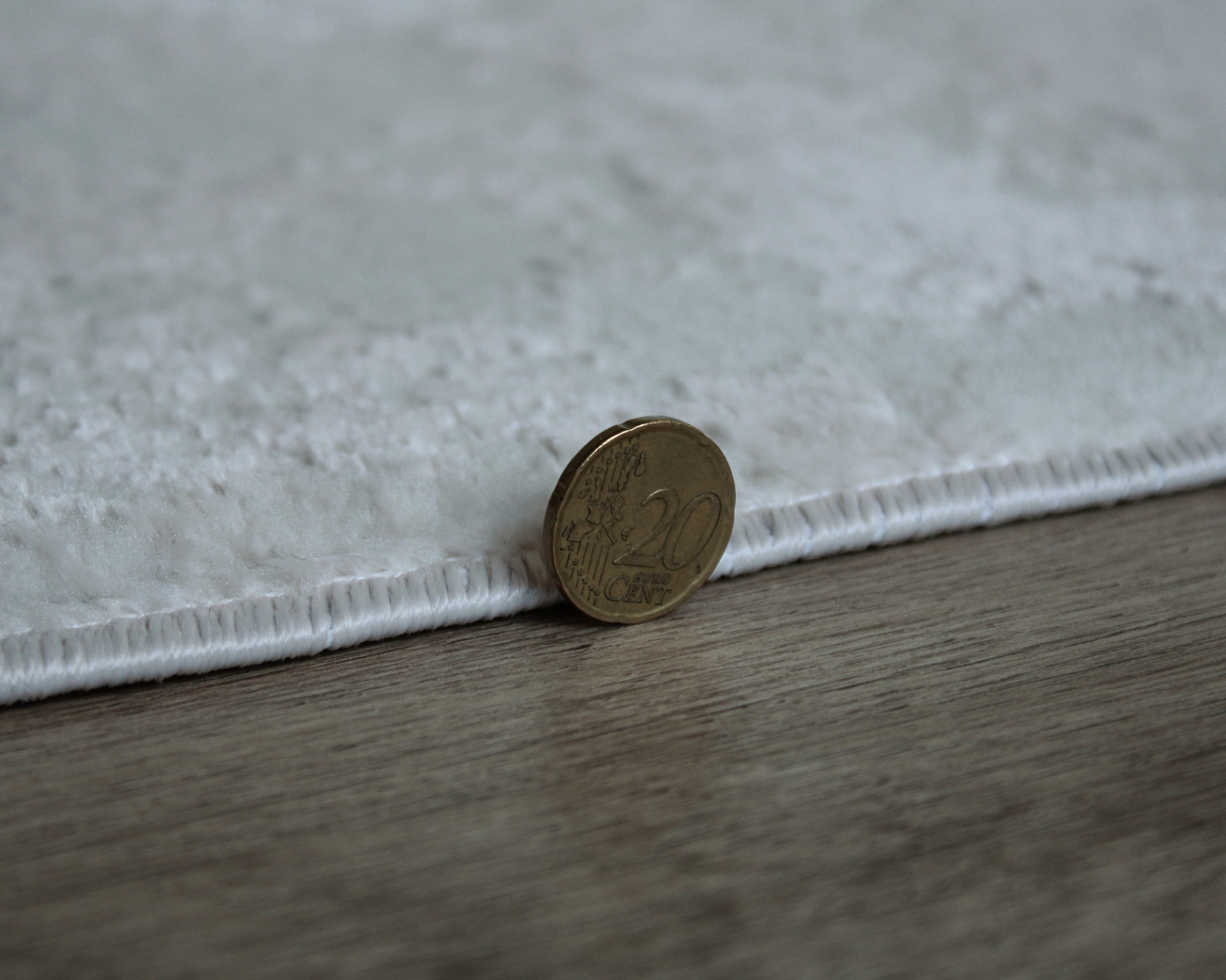 Tapete branco felpudo com moeda de 20 cêntimos encostada para comparação de espessura sobre chão de madeira.