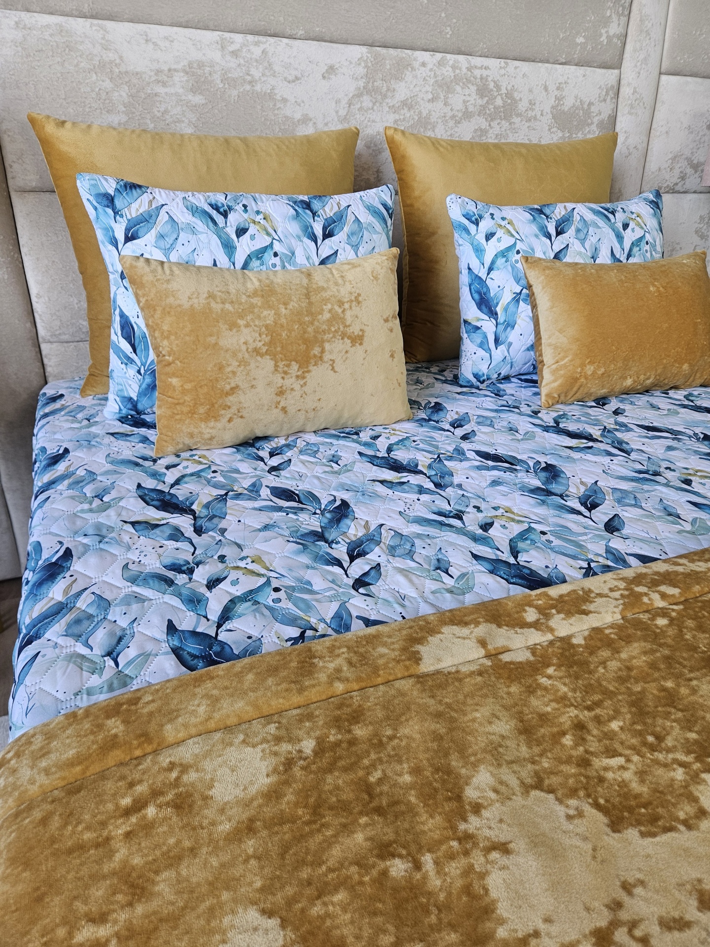Colcha e almofadas com padrão azul e veludo dourado numa cama com cabeceira cinza