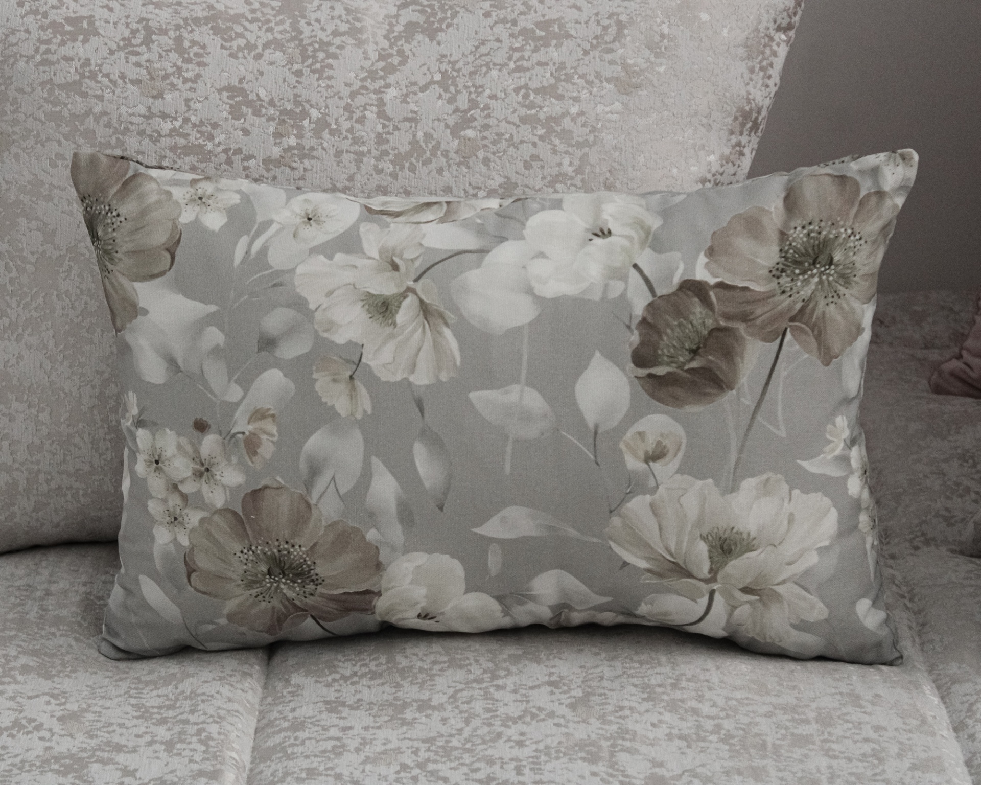 Almofada decorativa retangular cinza com flores sobre sofá bege claro.