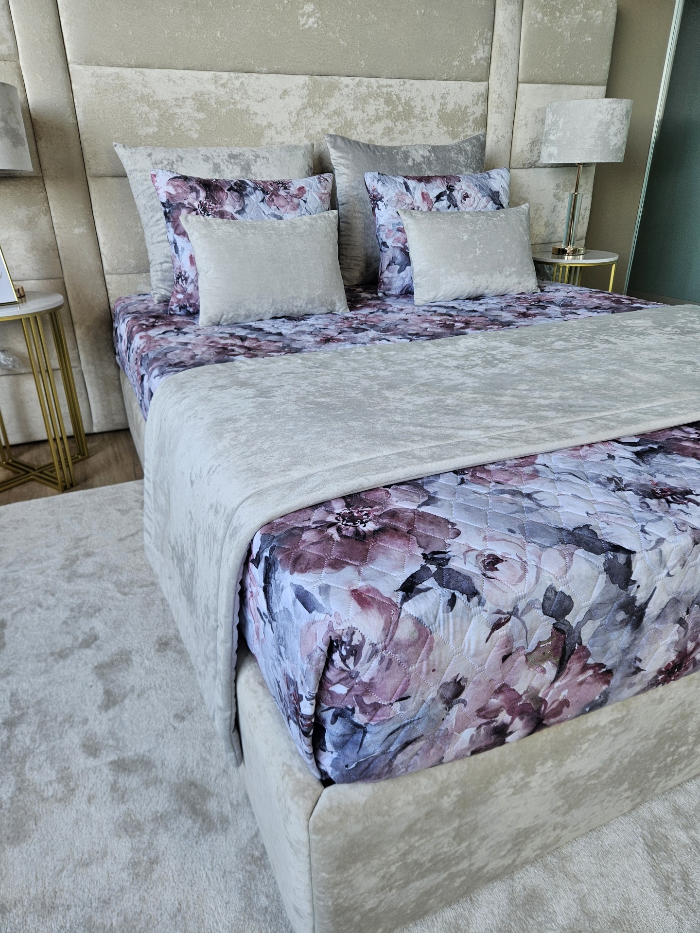 Cama decorada com jogo de cama acolchoado floral, almofadas cinza de veludo, cabeceira estofada cinza, mesas de cabeceira com candeeiros.