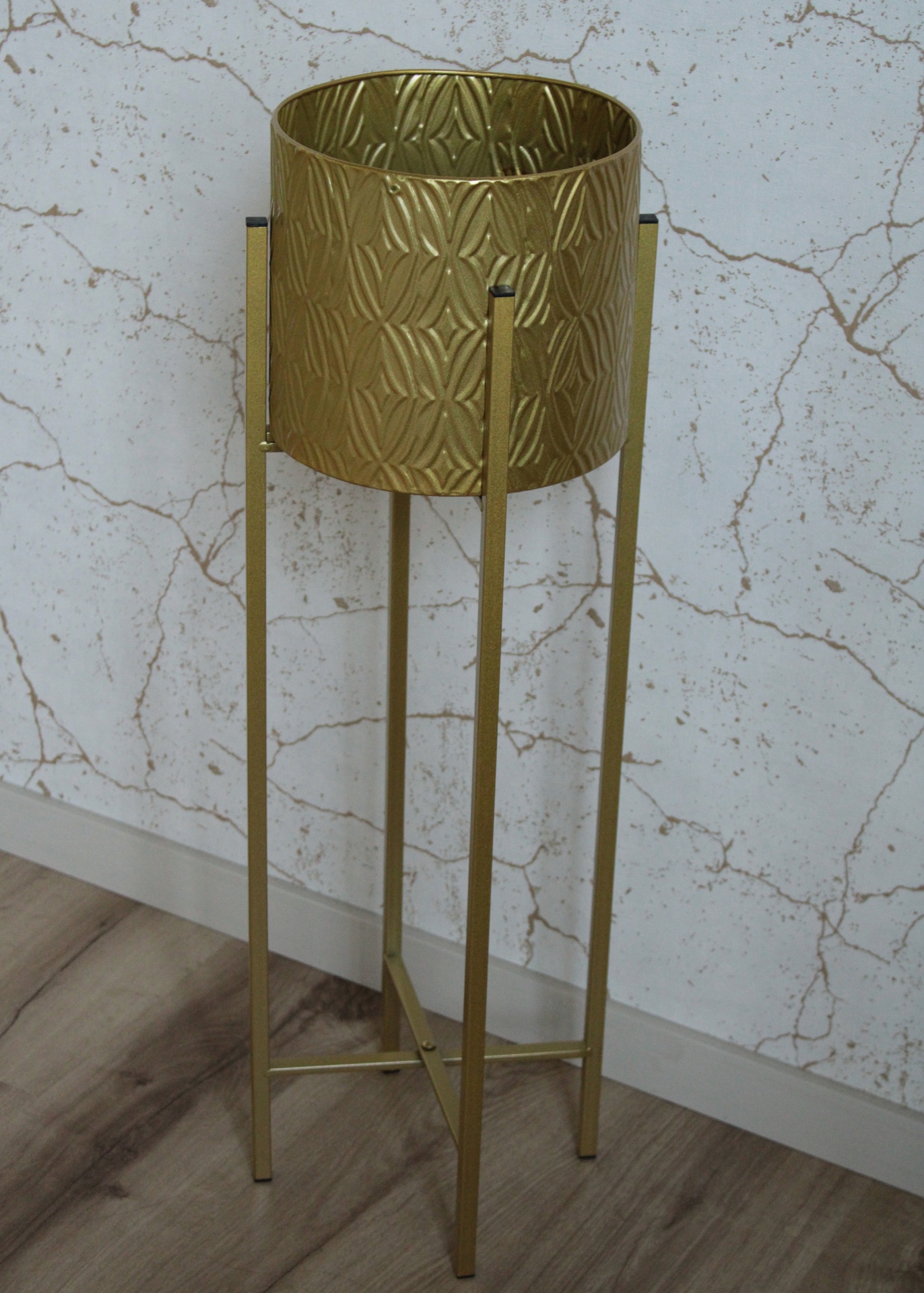 Suporte de planta dourado metálico com textura geométrica sobre chão de madeira clara e parede branca com padrão de rachaduras.