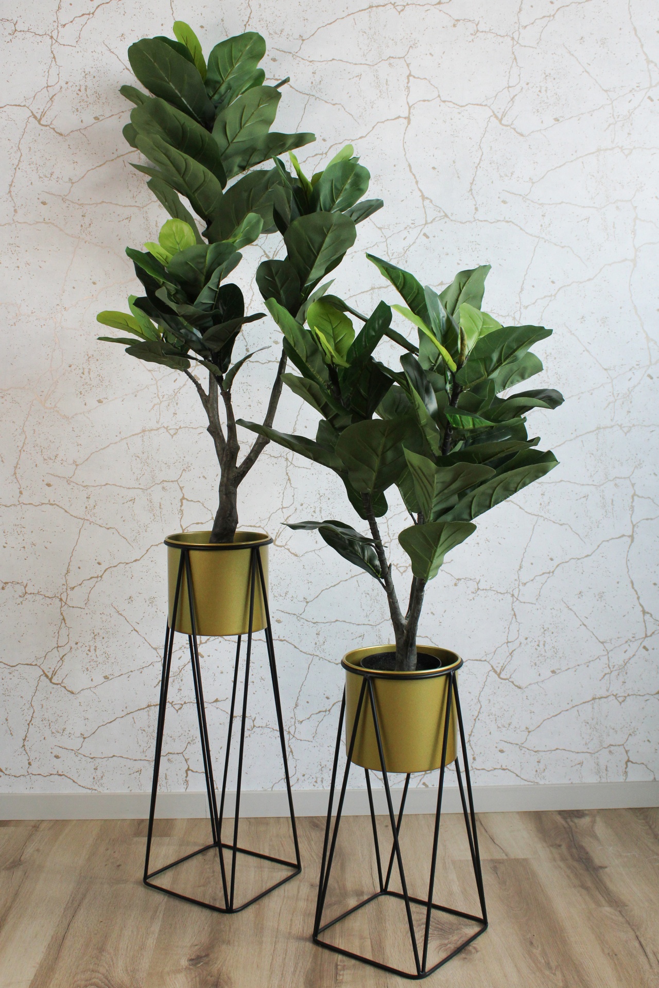 Dois vasos dourados com suportes pretos e plantas artificiais verdes em chão de madeira