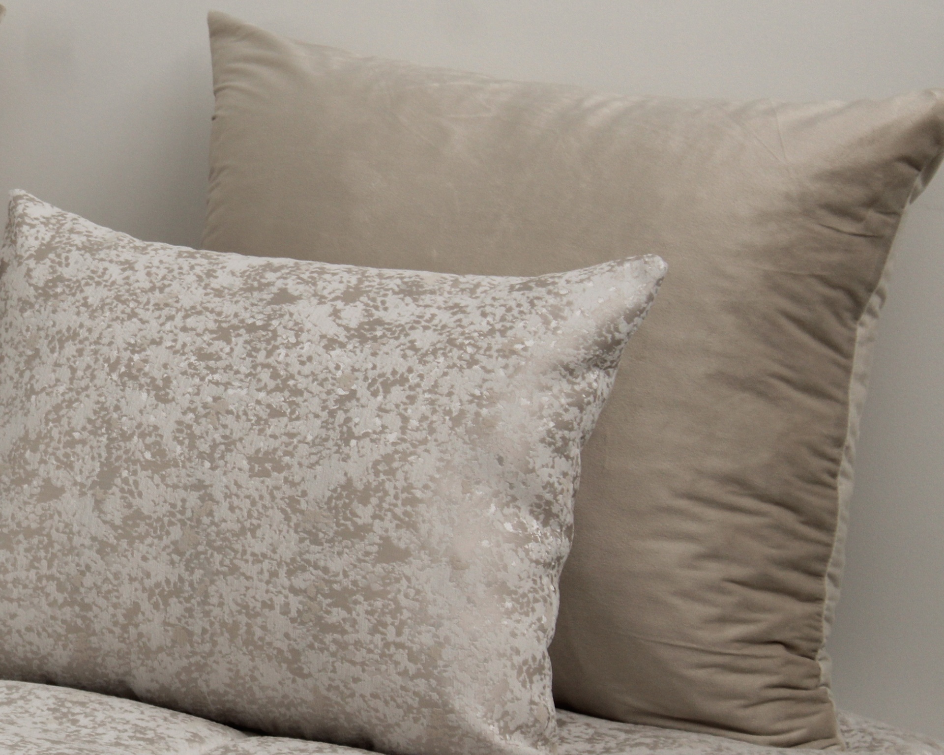 Duas almofadas, uma bege lisa e outra cinza claro com padrão abstrato, contra parede branca