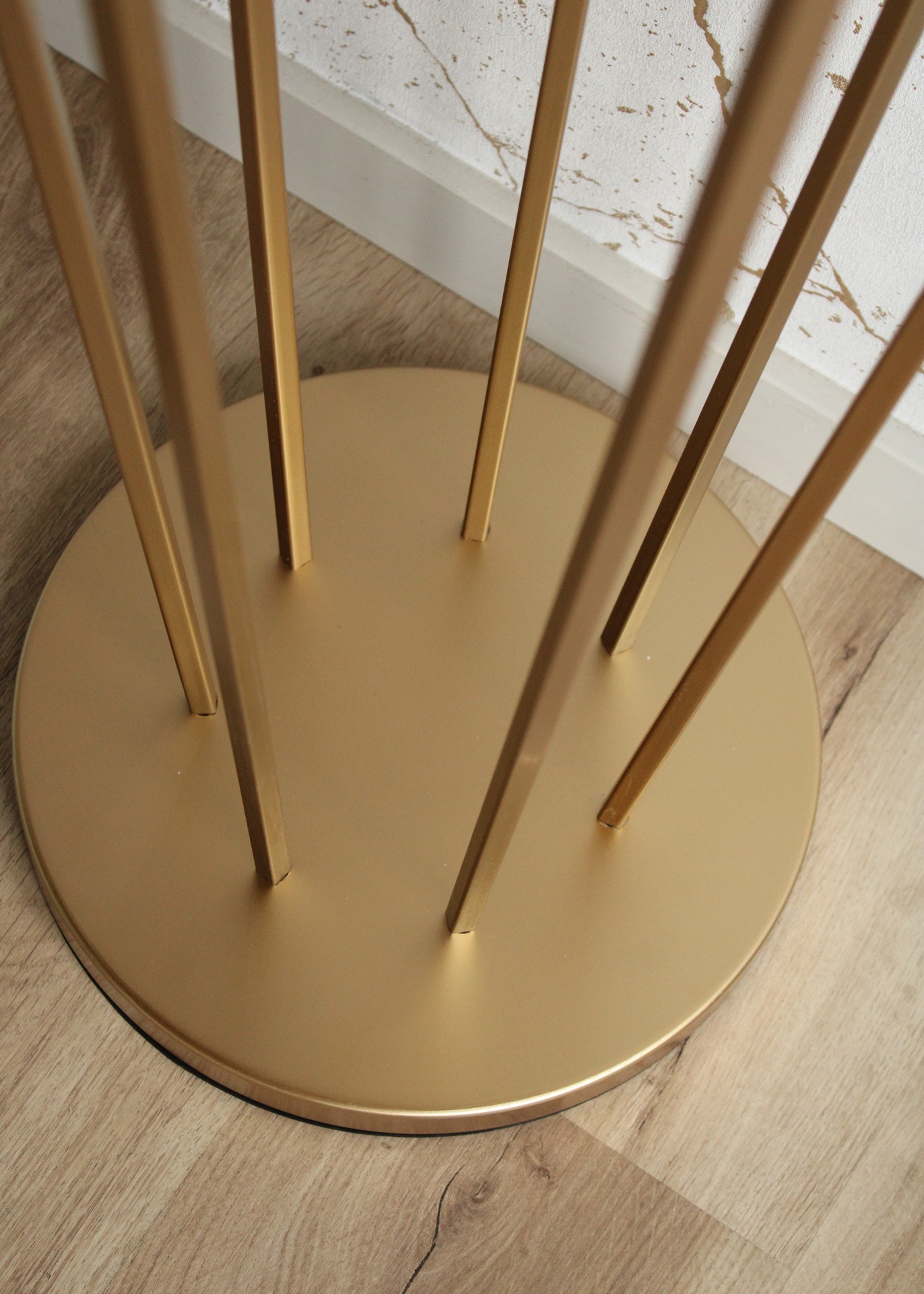 Base redonda dourada com hastes verticais em chão de madeira clara próximo a parede