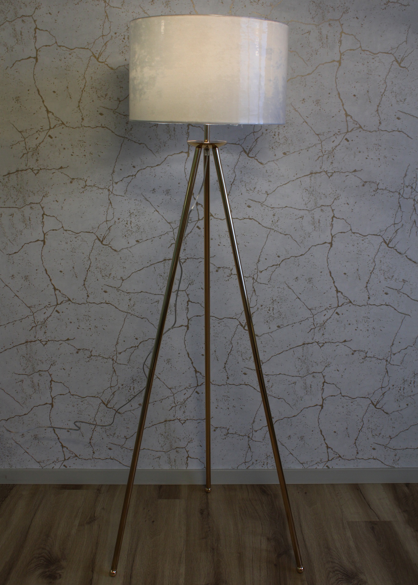 Candeeiro de pé com base metálica dourada e abajur branco frente a parede cinza