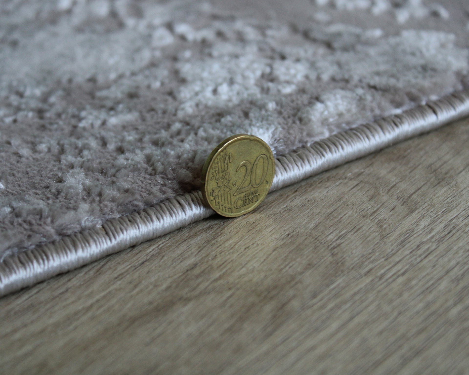 Moeda de 20 cêntimos junto à borda de tapete cinzento num chão de madeira.